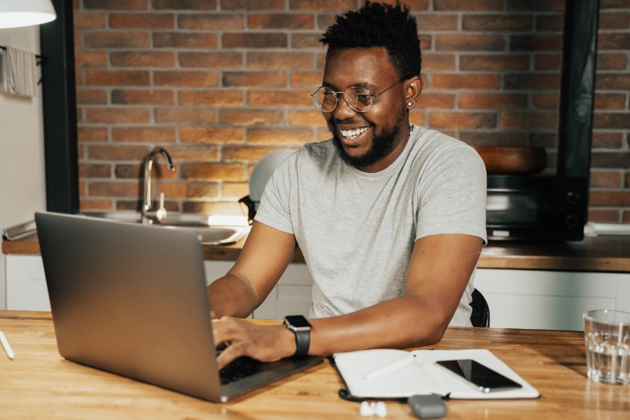 Homem negro trabalhando em um laptop prateado. Ele está sorrindo enquanto digita. Veste uma camisa cinza. Na mesa, está um caderno e um celular de tela escura. A parede atrás do homem é de tijolos expostos.